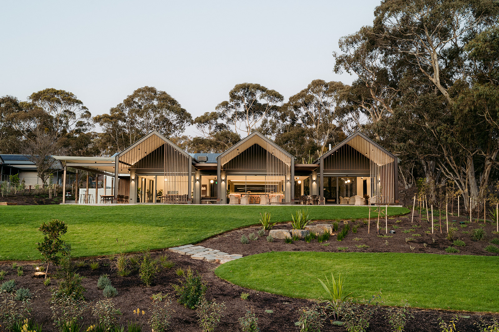 Three timber-battened gable pavilions with covered terraces opening onto a lawn, gum trees behind