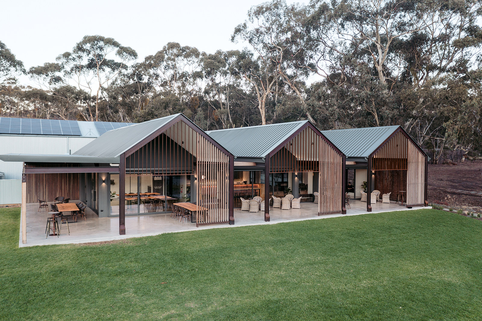 Three timber-battened gable pavilions open onto a lawn, with dining tables under the verandah and gum trees behind